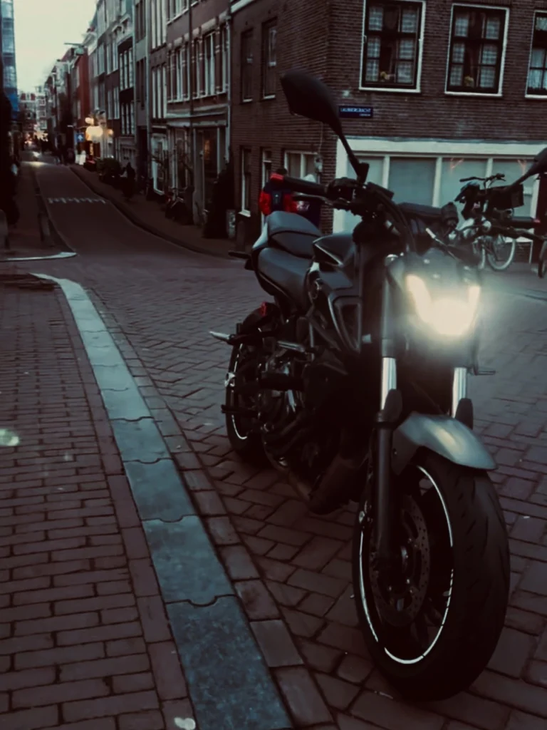 Motorcycle parked on an Amsterdam street at dusk with the headlight on